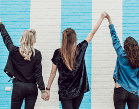Three women holding hands in front of a colorful wall.